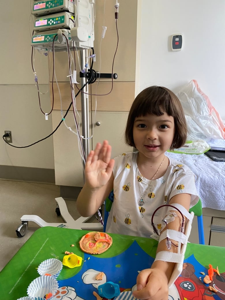 A little girl waves with her right hand. Her left arm is hooked up to a machine in a hospital setting.