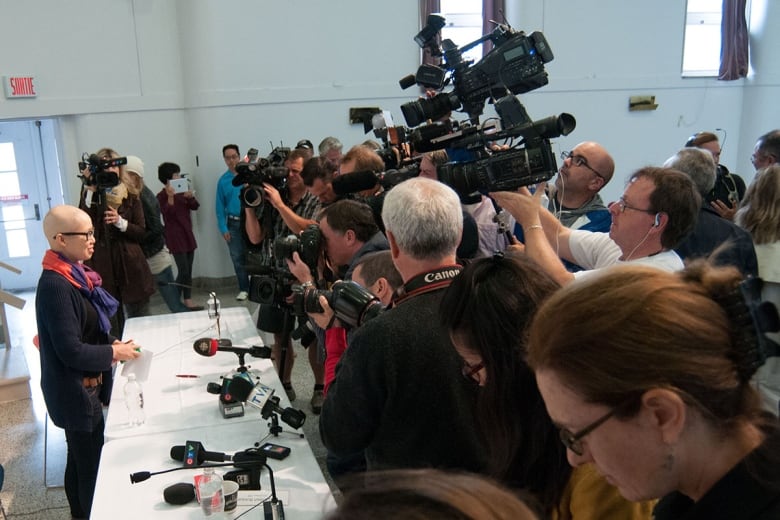 A small woman with a bald head stands behind a table. Facing her is a wall of TV cameras and journalists.