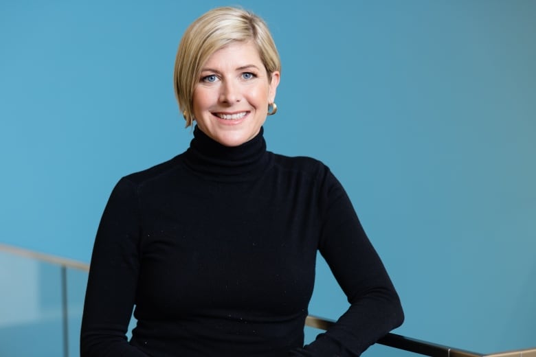 A blonde woman is seated in front of a blue background in a corporate portrait.