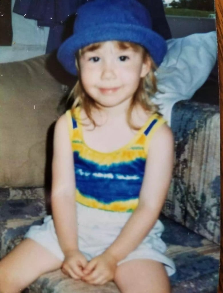 A little girl in a blue hat smiles at the camera. The photo is a scan of a physical copy and looks as though it was taken in the 90s. 