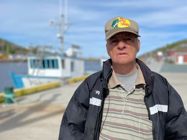 A man wearing a baseball cap and jacket stands on a wharf. 
