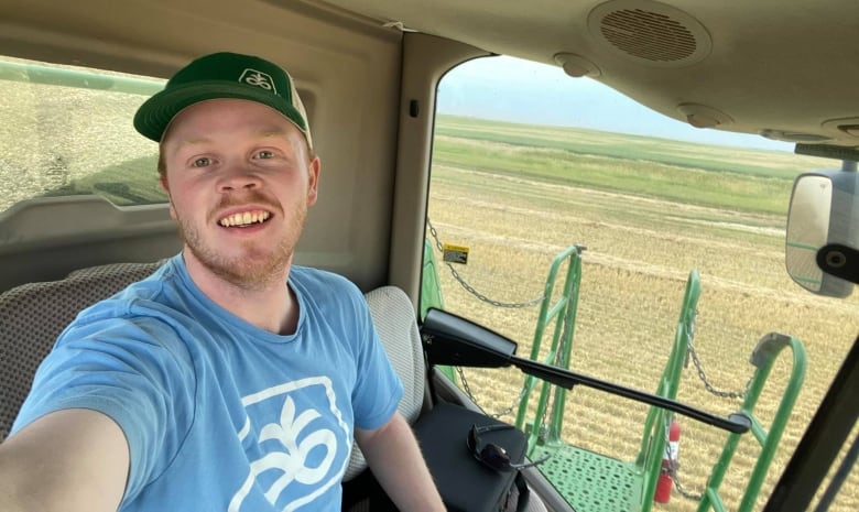 A man wearing a green hat and blue T-shirt sits in the cab of a farm machine with fields of crops visible through the windows behind him.