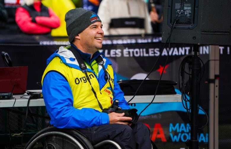 A middle-aged man in a toque sits outside in a wheelchair, a camera in his lap.