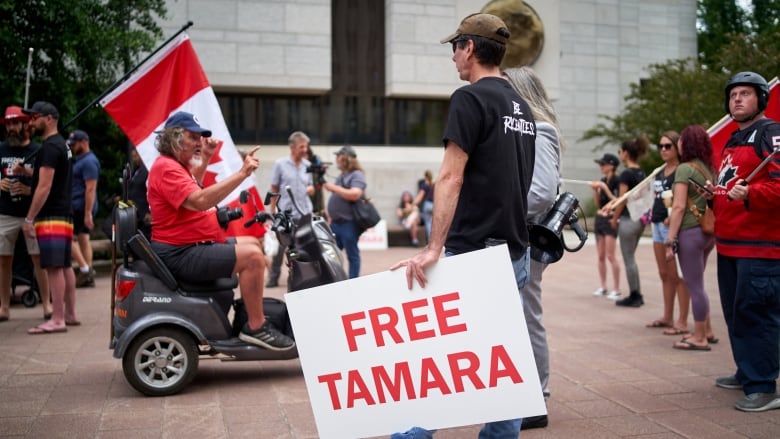 A man on a mobility scooter drives past a younger man in a black T-shirt holding a sign that reads "Free Tamara" in red letters.