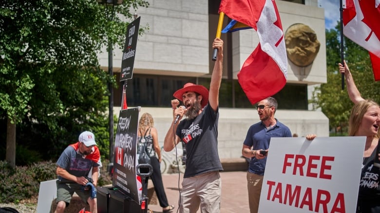 A man in a red cowboy hat shouts into a microphone while waving a Canadian flag. A woman holds a sign that reads "Free Tamara" in red letters.