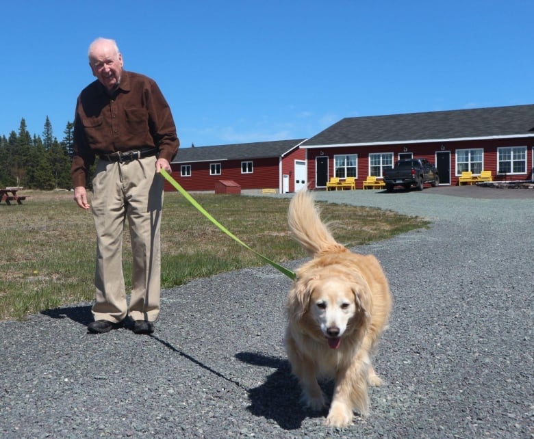 A smiling man walks a dog on a gravel path connected to a red building in the background. 
