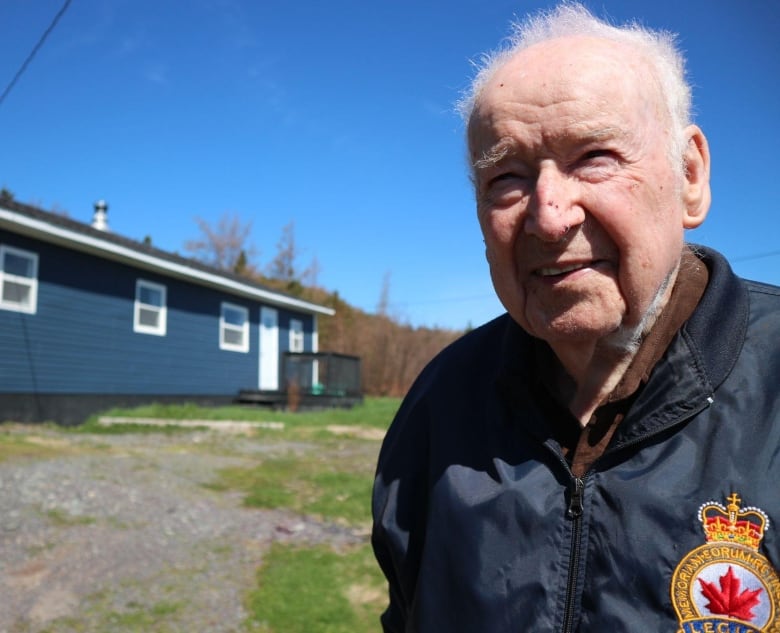 A smiling elderly man stands with a blue house in the background 