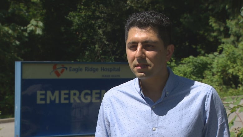 Ali Abdalvand looks at the camera. He is an Asian man wearing a blue spotted shirt. He is standing in front of a sign that reads 'Eagle Ridge Hospital Emergency'.