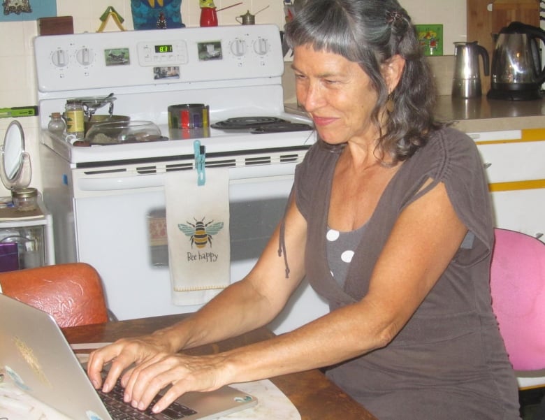 A woman sits at a kitchen table, typing on a laptop.