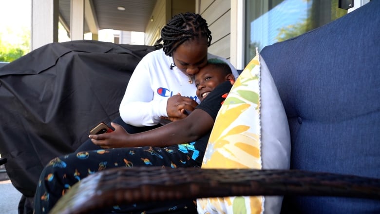 Jhody Batiste hugs her son on a porch. She is wearing a long-sleeved Champion sweater, he is wearing a black t-shirt.