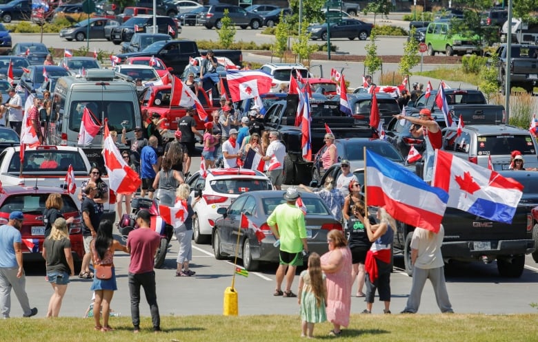 People gather in a parking lot, waving Canadian and Dutch flags.