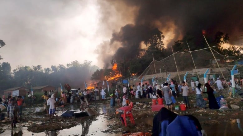 A crowd of people in the foreground with flames and smoke rising above structures in the background. 