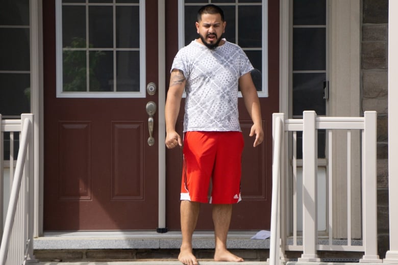 A barefoot man wearing a grey t-shirt and red shorts stands on the steps of a home. 