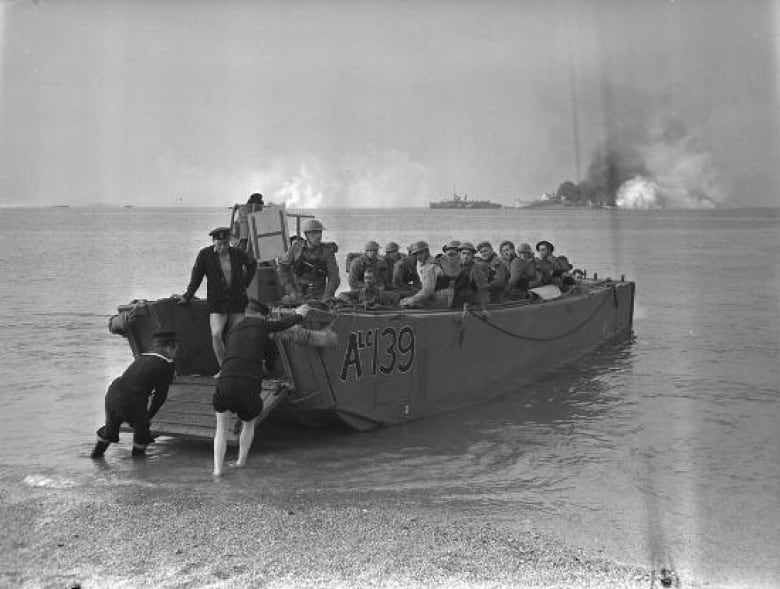 A boat full of men in military uniforms is being pushed off the beach by two others who are up to their ankles in the water. In the distance, there are ships and plumes of smoke. 