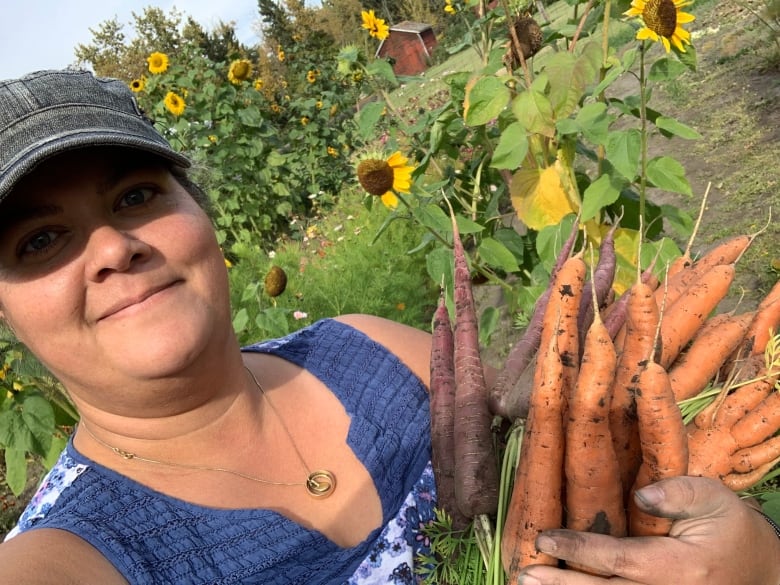 A woman holds up an armful of carrots in a garden of sunflowers.