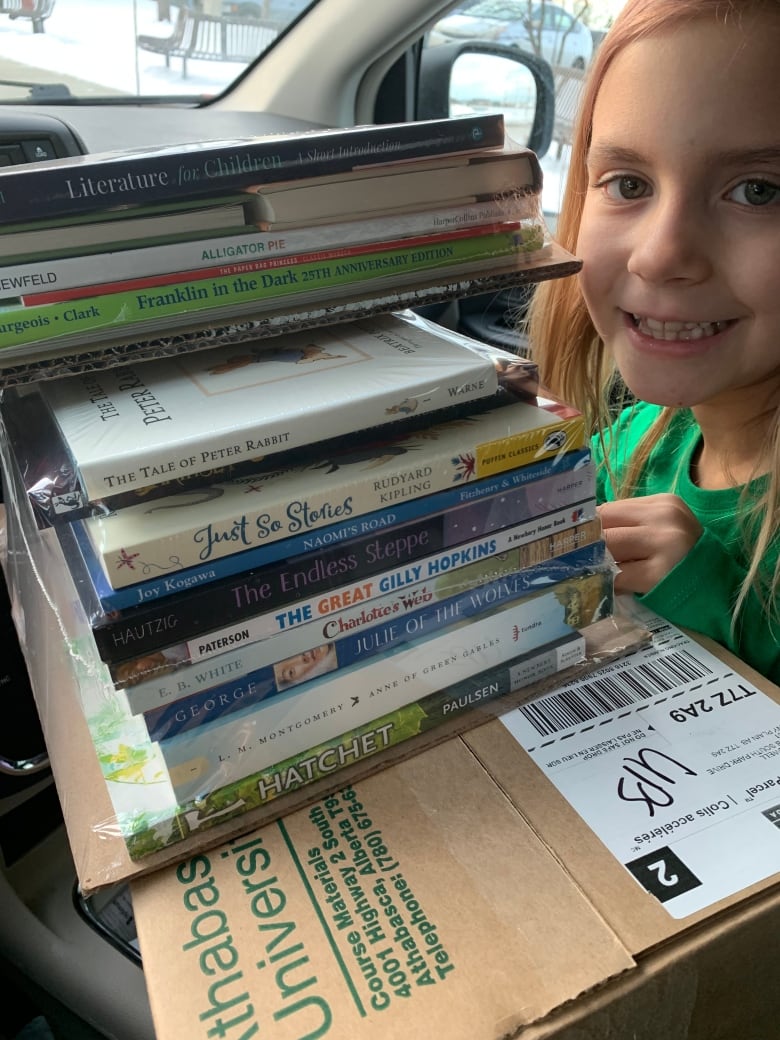 A smiling girl stands next to a stack of children's books and university textbooks.