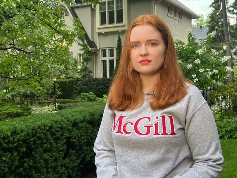 A young woman with long red hair and wearing a grey McGill University sweatshirt stands in a front garden outside a house.