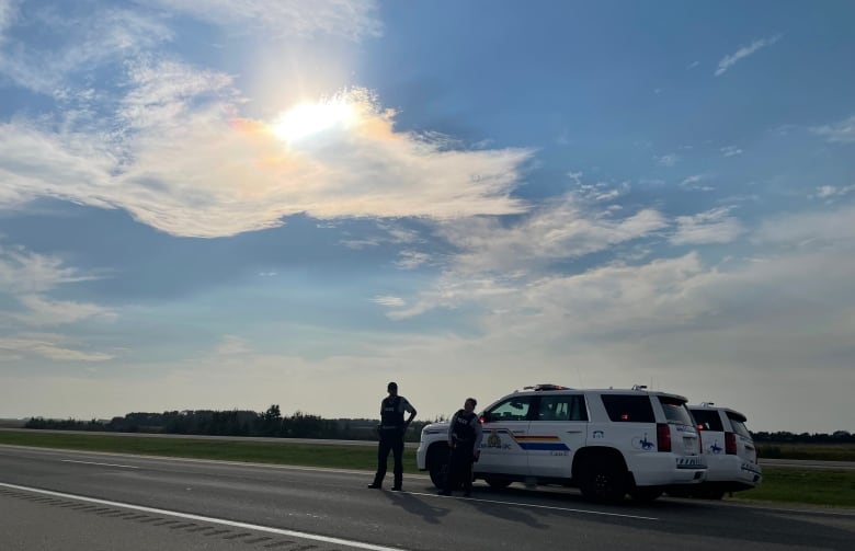 Two RCMP officers stand near two RCMP vehicles on a highway under a partially coudly sky.