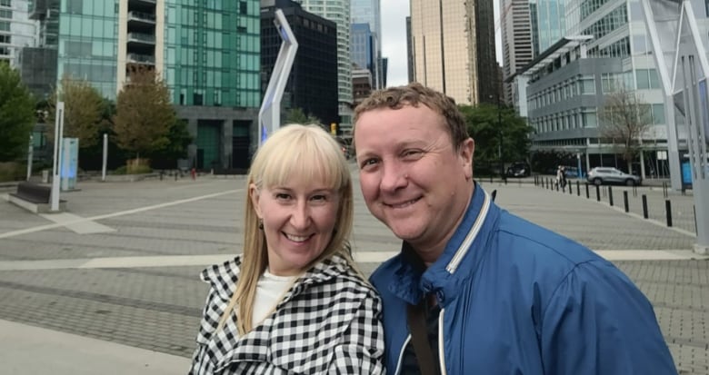Two people, a husband and wife, smile in front of an installation. The wife has long blonde hair and is wearing a black-and-white checked top. The husband is a white man with short brown hair, wearing a blue jacket.