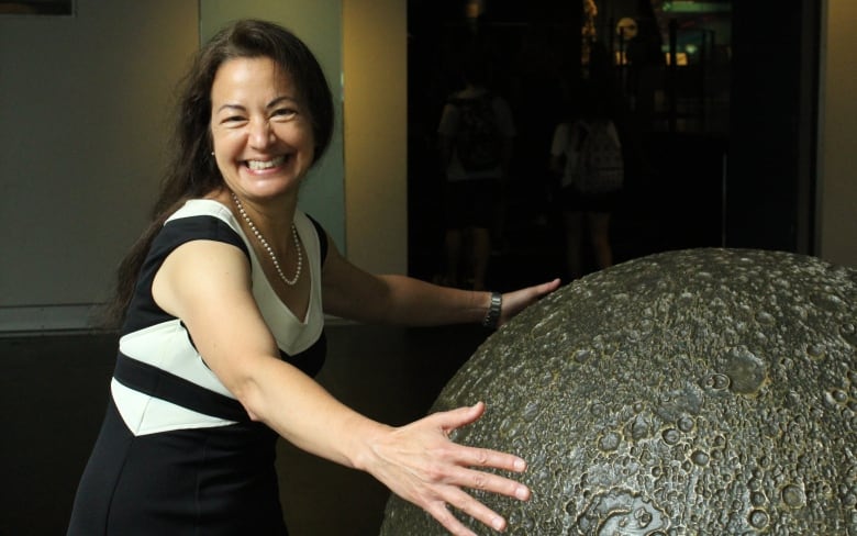 A woman in a black and white dress smiles, while embracing a model of the moon.