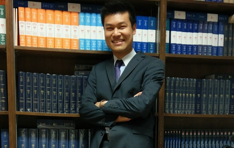 A man in a dark suit stands, smiling, in front of a bookshelf.