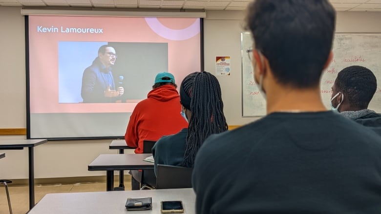Students are seen from behind as they listen to a speaker projected onto a screen at the front of the classroom.