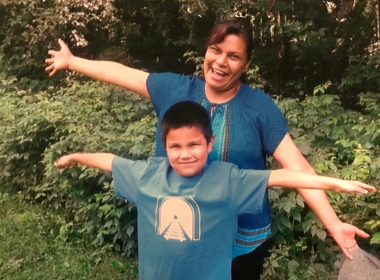 A woman and a little boy smile while spreading their arms out wide, in front of a green bush.
