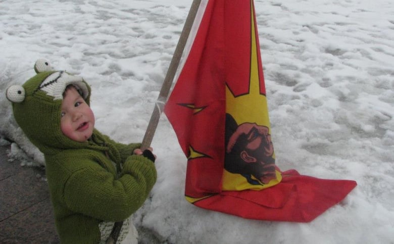 A toddler in a green frog jacket holds a red flag on snow.