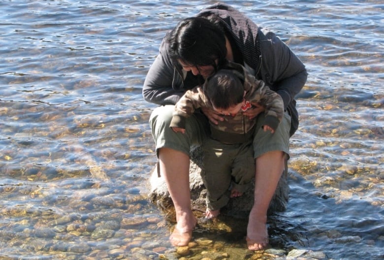 A woman holds a baby boy and they both look down as she stands in a shallow lake water.