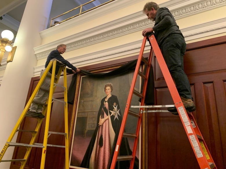 Two men on ladders hang a black cloth on a portrait of a woman wearing a long robe and a crown on her hand. 