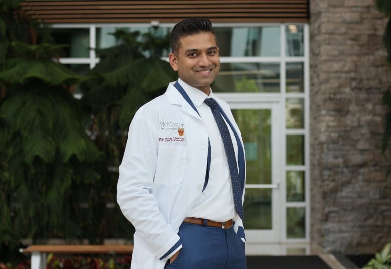 An Indian man wearing a suit, tie and lab coat stands in front of a building. 