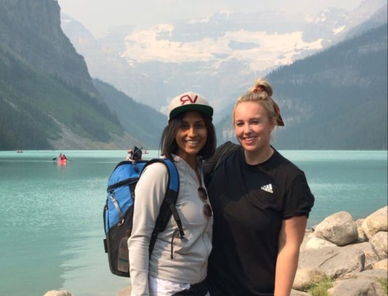 Two smiling woman stand in front of a lake with the mountains in the background.