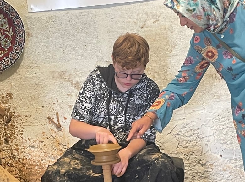 A woman in a vibrant headscarf and dress guides a teenage boy as he shapes a small clay bowl in a pottery demonstration.