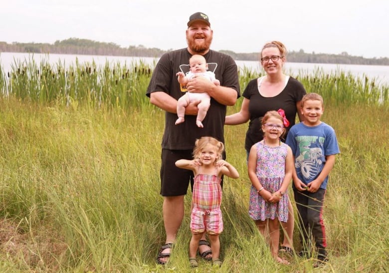 A smiling family of six -- mom, dad and four children ranging in age from six months old to eight years old -- stands outdoors in a tall grassy field next to a waterway.