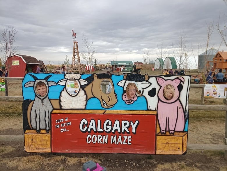 Three children make funny faces as they pose behind a photo cut-out board, their faces peering out from a painting of a goat, a cow and a pig. Farm buildings and structures are seen in the background. 