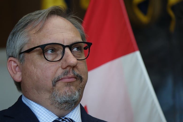 A man in a suit listens during a press conference. he wears black glasses and is standing in front of a Canadian flag.