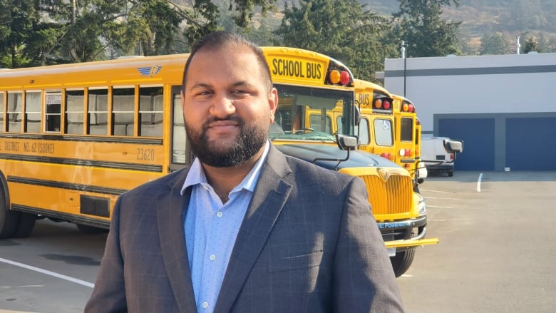 A bearded man wearing a grey suit and blue shirt stands outdoors in a bus parking lot, in front of several yellow electric school buses. 