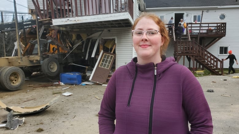 A woman looks into the camera, with a destroyed building behind her.