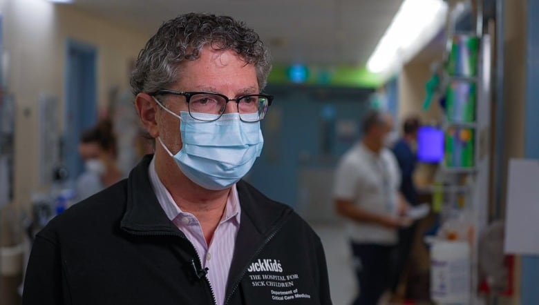 Man in a blue surgical or procedure mask stands wearing a SickKids sweater.