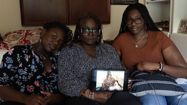 Two teenagers and one woman sit on a couch, holding a photo of another woman and a baby. 