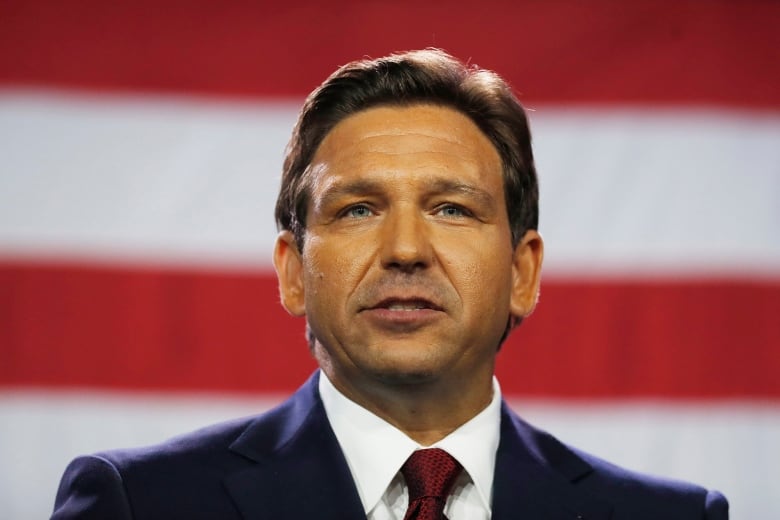 A man with short brown hair, wearing a navy blue suit with a red tie, visible from the shoulders up, in front of the red and white stripes of a large American flag.