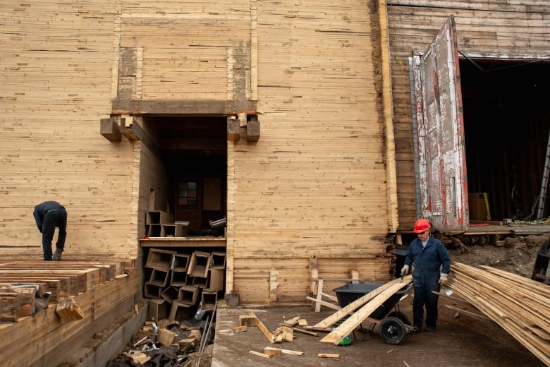 Two men work in front of a grain elevator that has been stripped of its siding, exposing the wood underneath. They are piling the planks in front of it.