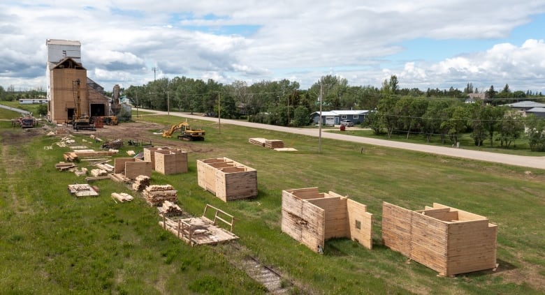Piles of wood and box-like structures made of planks of wood sit organized in rows next to a grain elevator that is partially demolished.
