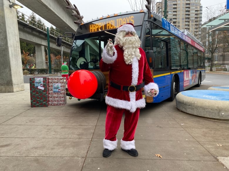 A transit bus is pictured decorated with reindeer ears and a nose. Santa Clause stands in front handing out candy canes. 