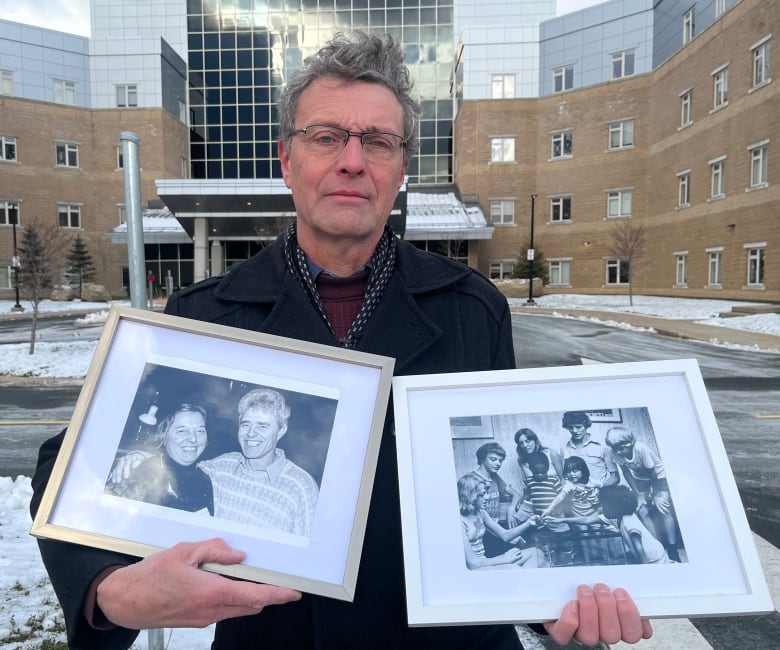 An older man is holding two framed black-and-white photographs. The one on the left is showing a young couple, the one on the right a family of eight. He stands in front of a big, four-storey building.