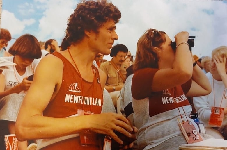 A photo shows a man, left, and a woman, right, who are looking to the side, at something off camera. The woman is looking through binoculars. The man is wearing running attire. Both their tops read "Newfoundland". Behind them, there are rows of people. People's attire and hairstyles show that the photo was taken decades ago.
