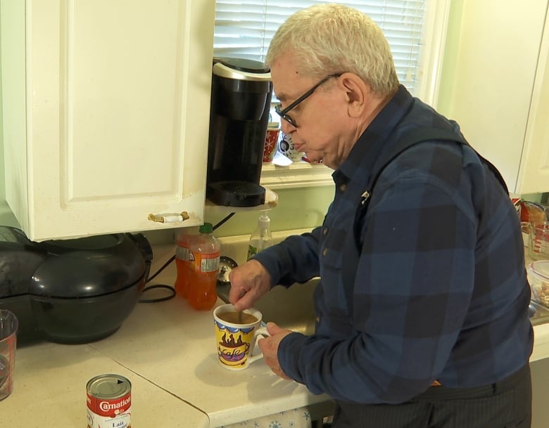 An older man, on the right-hand side of the photo, is standing at a kitchen counter, stirring a cup of coffee.