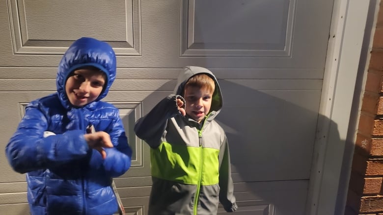 Two little boys wearing winter jackets stand in front of a garage door. Both are giving a thumbs down. 