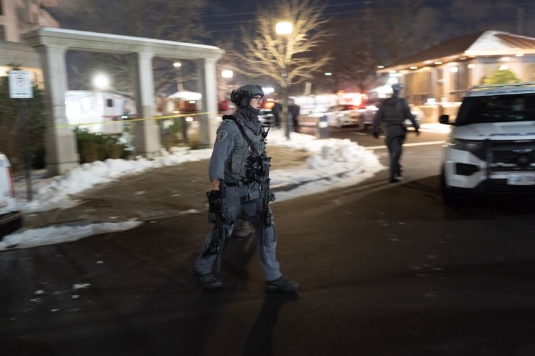 A police officer, in a grey uniform and helmet, crosses a road outside an apartment building at night.