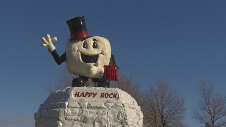 A picture of a famous roadside attraction in Gladstone. It is a rock with a face on it and it is smiling and waving and the words Happy Rock under. The rock is wearing a black top hat. 